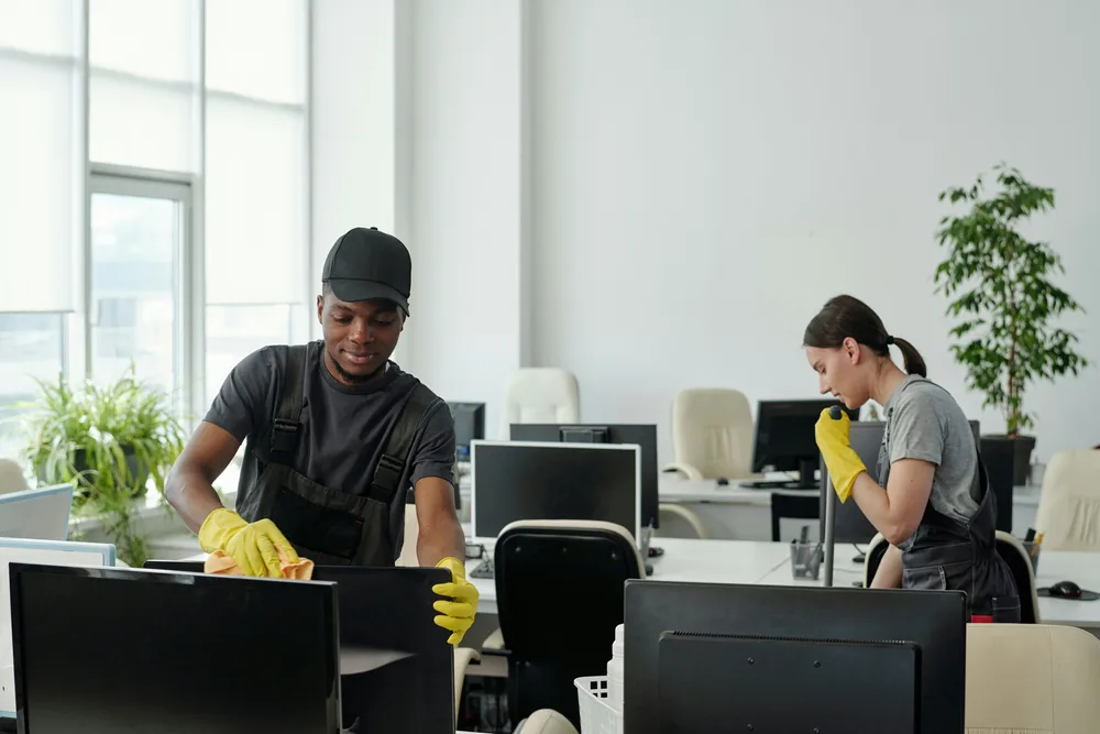 Two cleaners wiping desks and computer equipment in a bright office